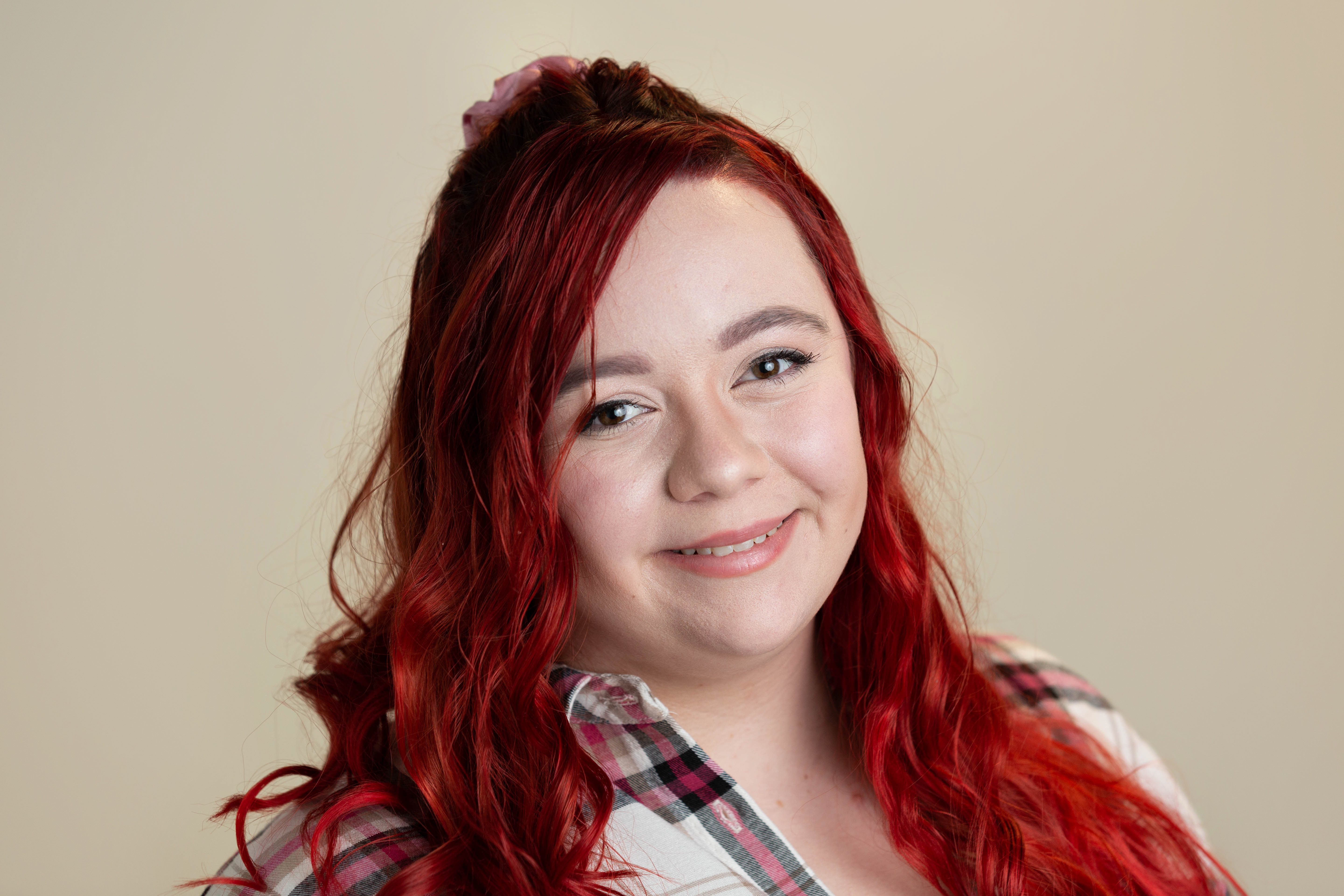 A white woman with long red hair smiles at the camera, wearing a checked shirt