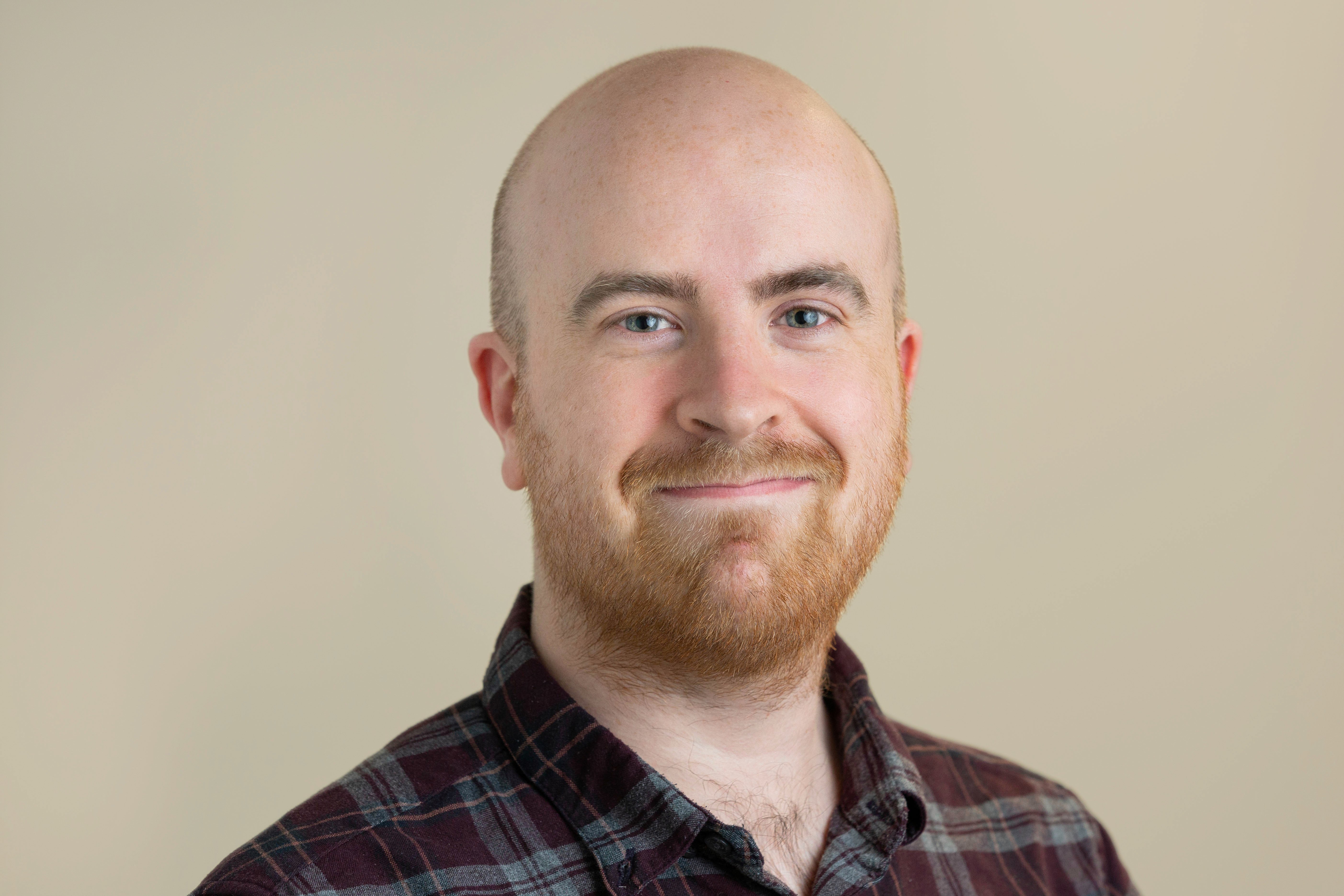 A white man smiles at the camera, wearing a checked shirt 