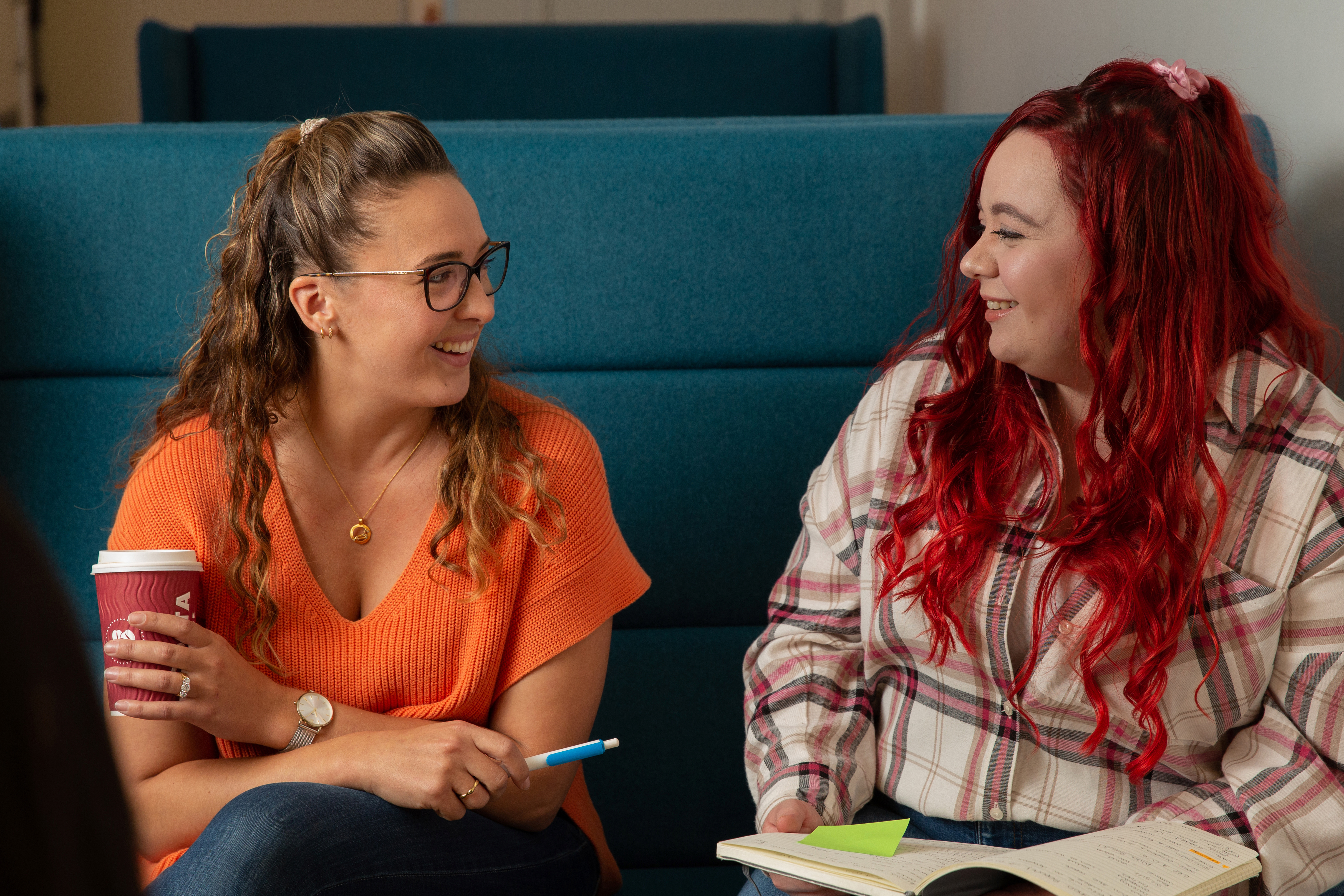 Two women smile at each other while sat on a sofa, holding a coffee cup