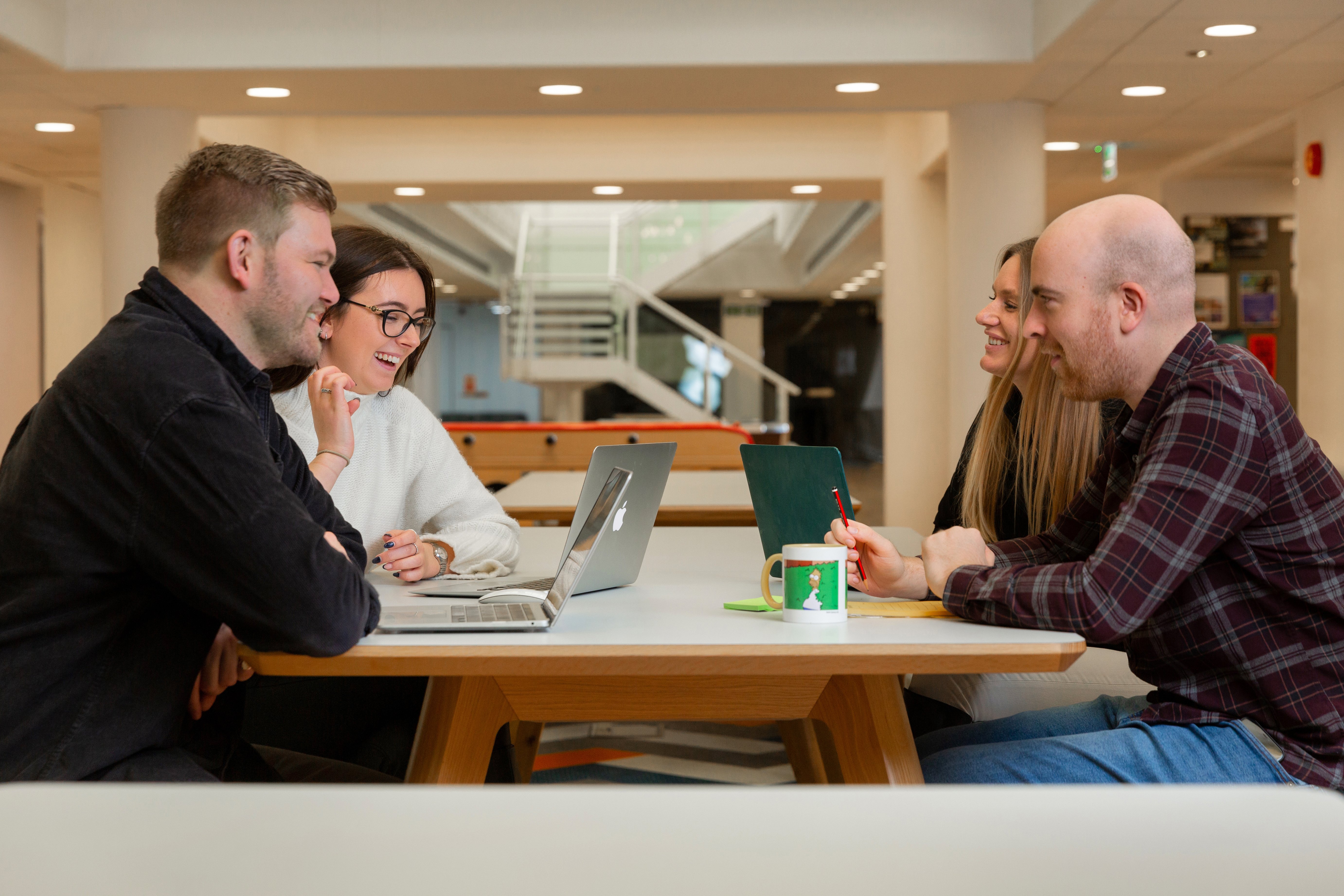 A team of people are talking in a meeting around a table, one person is laughing and everyone is on a laptop.